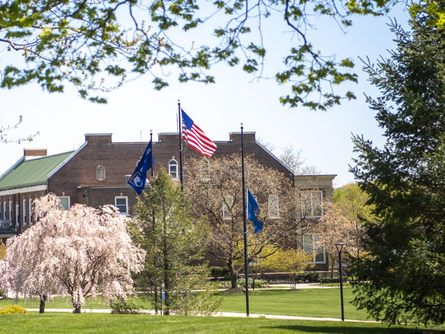 U.S., Pennsylvania, and Penn State Schuylkill flags fly in the wind over a large grassy field with a brick building in the background.