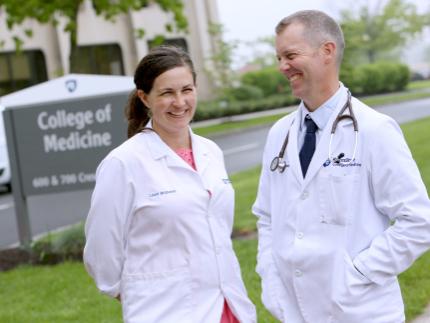 Two people – a man and a woman – stand side-by-side wearing white physician’s coats. They’re on a lawn in front of a large building, with a “College of Medicine” sign in the background.