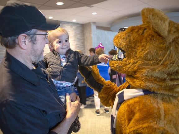 A man holds his young grandson as they laugh at a man dressed in a lion costume