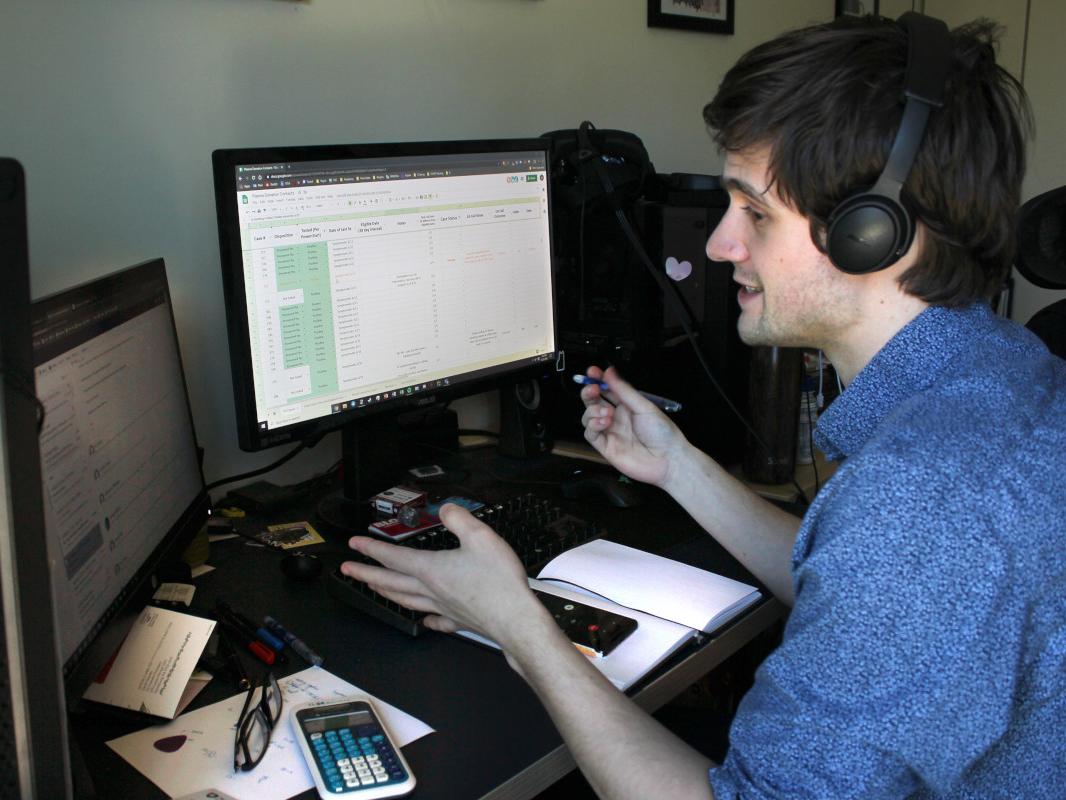 A man sits at a desk in front of two computer monitors. He's wearing headphones and holding a pen in his right hand. A calculator, eyeglasses and other items are on the desk.
