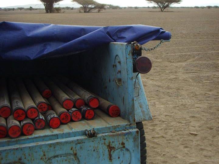 Light blue open-backed truck with drilling cores. African arid landscape with sparse trees in the background.