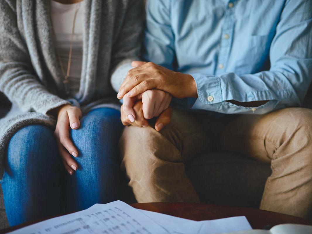 A man and a woman sit side-by-side on a couch. Image shows them from the neck down. The woman’s left hand is on the man’s right leg, which he holds with both hands.
