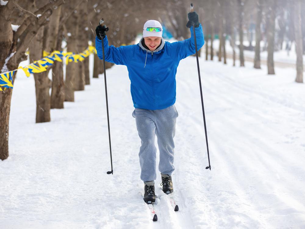 A man wearing winter clothing uses cross-country skis to make his way along a tree-lined road that is coated in a few inches of snow.