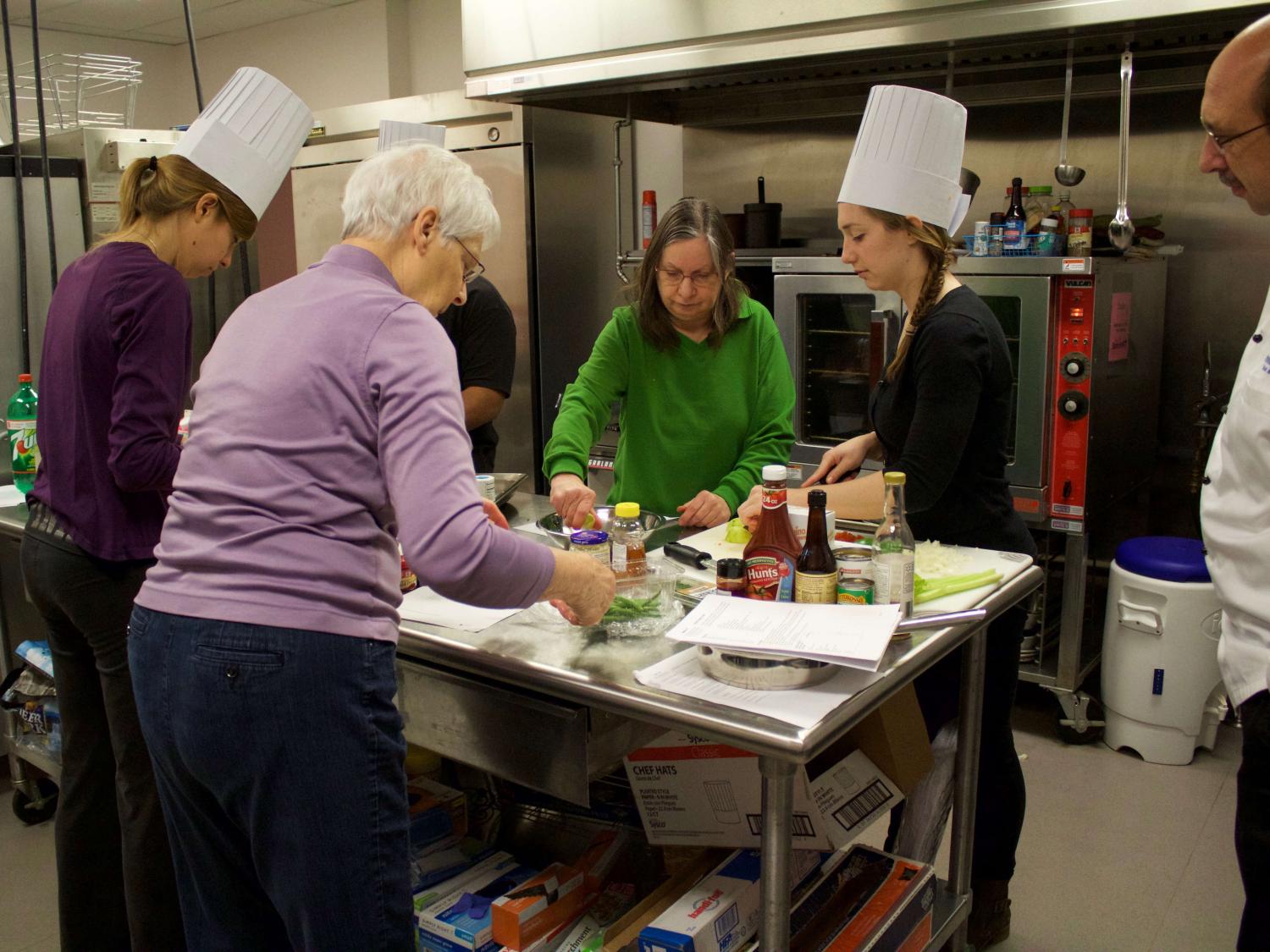 Four people prepare food on a table in the middle of a large kitchen while one person looks on.