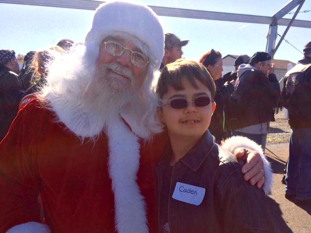 Santa Claus and a child with a 'Caden' nametag pose for a photo. Motorcyclists walk around in the background.