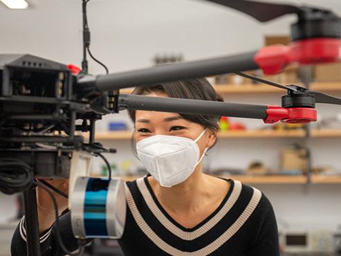 A woman in a black sweater and a white face mask adjusts a black drone.