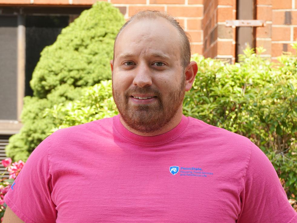 Smiling man in a pink t-shirt standing outside a brick building with green bushes behind him.