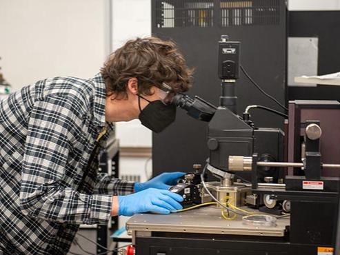 A person wearing a black face mask, blue gloves and a plaid shirt looks into a microscope. 