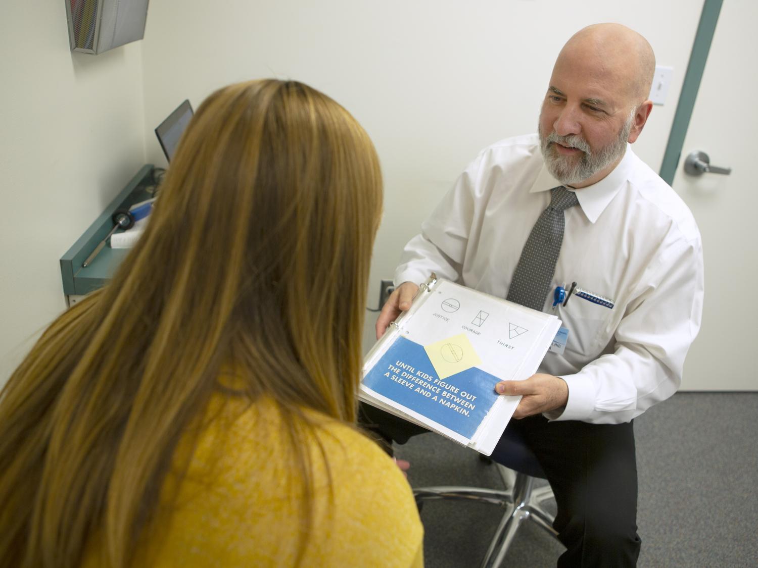 A male physician wearing a tie sits on a stool in an exam room, displaying a page with various diagrams on it to a female patient who sits across from him.