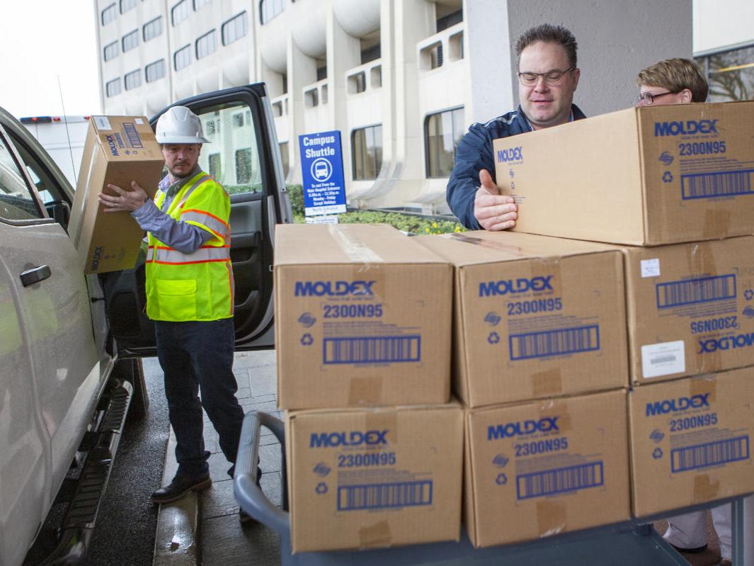 Three people -- two men and a woman -- load large boxes from a vehicle onto a cart. Hershey Medical Center is in the background.