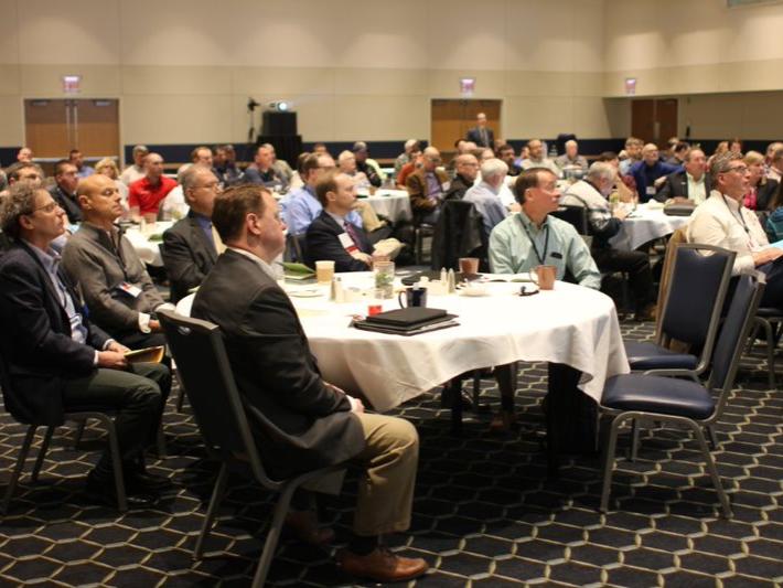 A room full of people sitting at round tables watching a presentation