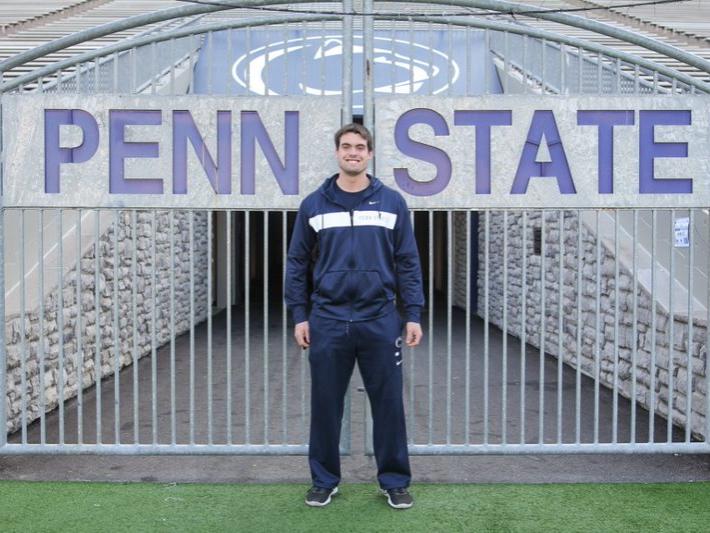 A student, wearing all blue, stands before iron gate that opens into the Penn State football field. The gate has the words Penn State across the center, with the Penn State Nittany Lion athletic logo looming behind it from the stands.