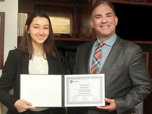 A woman in a suit jacket and a man in a suit smile and pose with a certificate