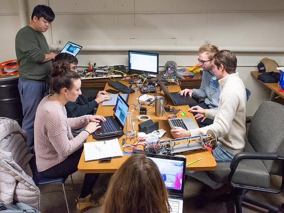 Six students sit with their laptops around a table, which is covered with wires and electronic components. 