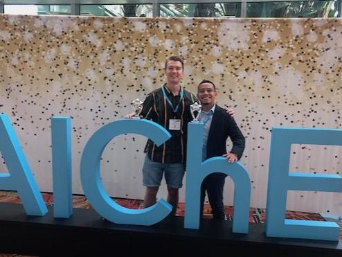 Two young men standing in front of large letters spelling out "AIChE"