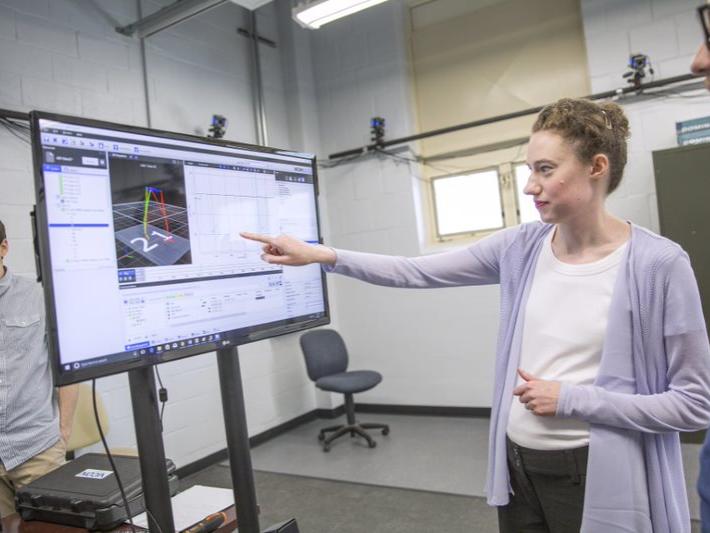 A Caucasian women points to a screen with computational modeling, with two Caucasian men in the background. 