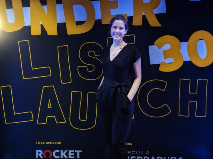 A Caucasian woman poses in front of signage that reads "Forbes 30 Under 30 Launch."