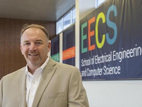 A man in a tan suit jacket smiles at the camera in front of a colorful EECS banner.