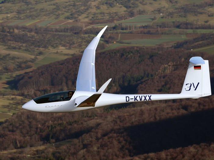 A smiling man gives a thumbs up from the cockpit of a small glider plane flying over forest. 