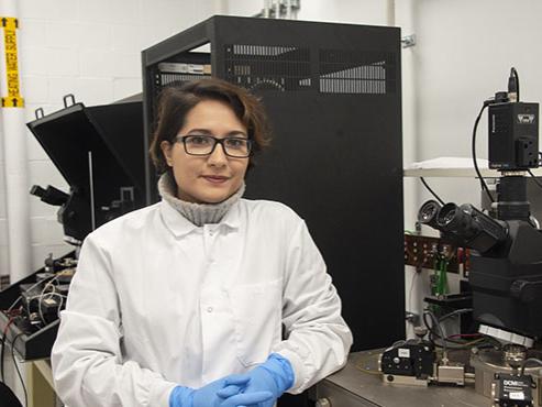 A woman in a white lab coat, blue gloves, and glasses sits next to a microscope in a lab and smiles at the camera 