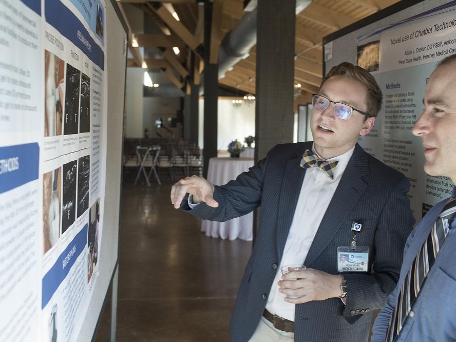 The poster at which Jason Spicher gestures is reflected in his glasses. He holds a glass in his other hand. Next to him, another man smiles at the poster. Behind them, support beams rise into the distance.