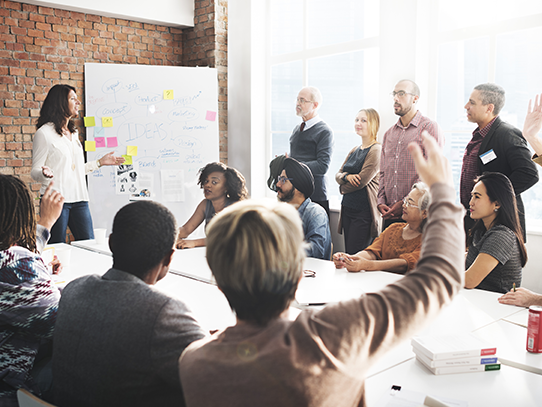  A dozen people sit and stand around a table as a woman leads a discussion at the front of the room. 