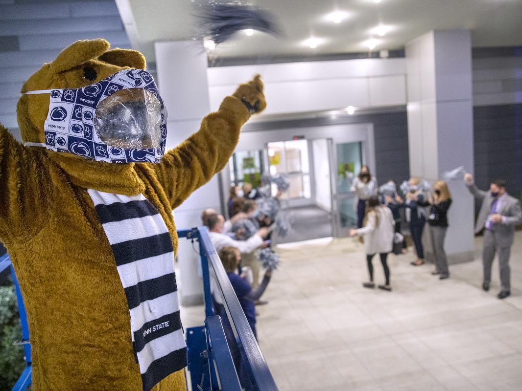 The Penn State Nittany Lion stands in the foreground, with several people in the background cheering as an individual walks through a doorway.