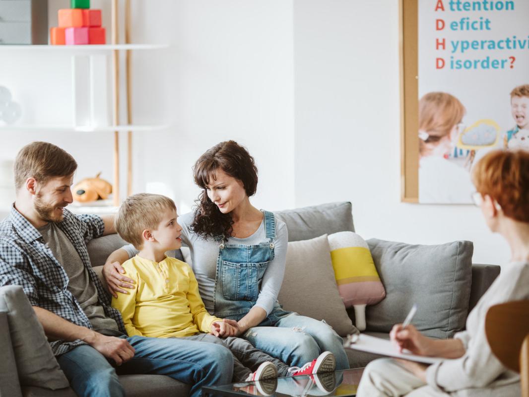A man, young boy and woman sit on a couch. The man and woman have an arm around the child, who is in the middle. A woman with a notepad sits across from them in a chair.