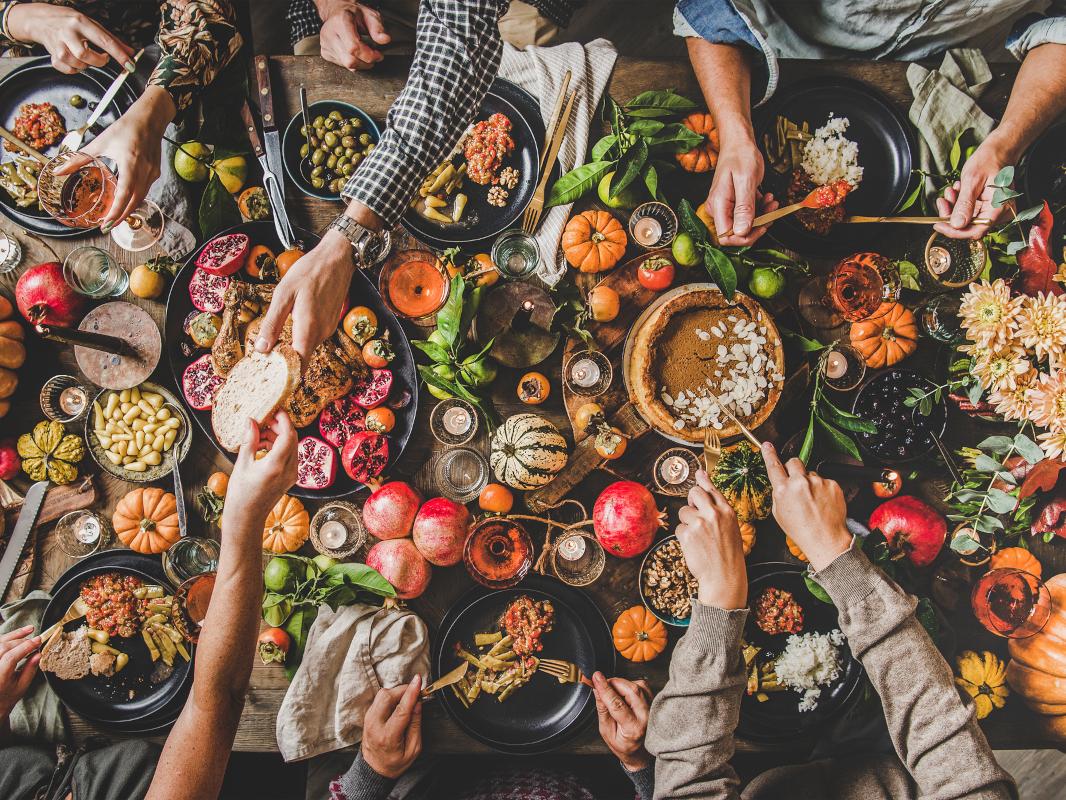 An overhead view of a table at which six people are seated. Their arms are stretched out over the table, some over their plates, others getting food. A wide range of bread, grains, fruits and other food is on the table.