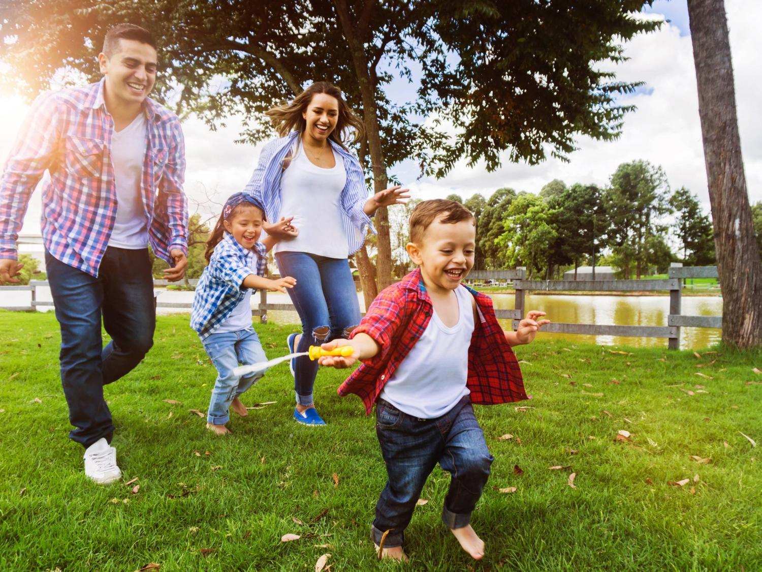 A man, a woman, and two children smile as they run through a park. trees and a fence are in the background.