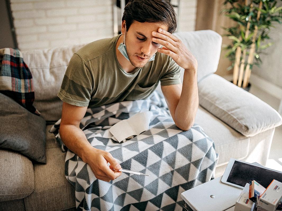 A sick man wearing a mask around his chin takes his temperature using a digital thermometer. 
