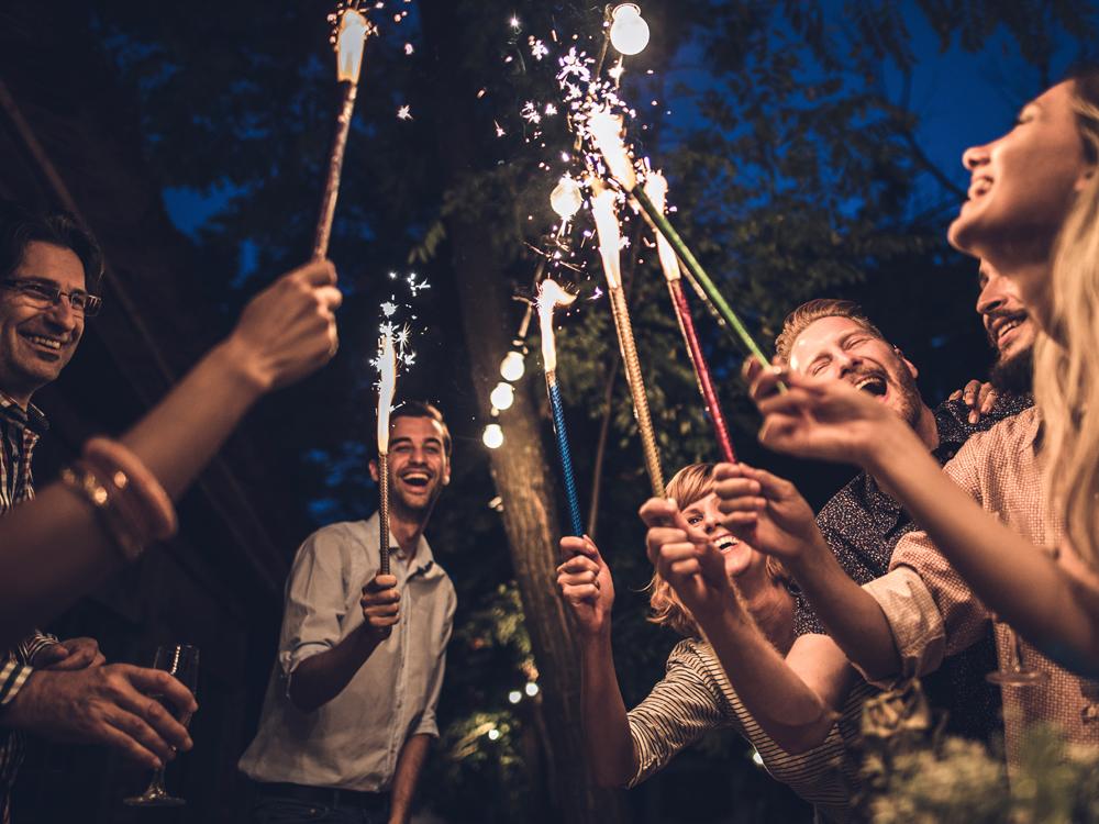 Several people stand in a circle, smiling, and holding sparklers toward the center of the circle. The vantage point is looking up from the ground.