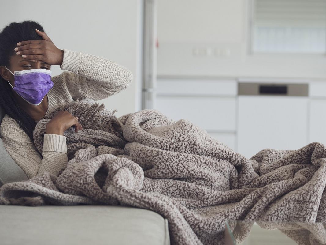 A woman lying on a couch under a blanket and wearing a surgical mask presses a palm to her forehead.