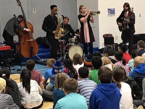 Musicians stand while they perform in front of a group of middle-school-aged students sitting on a floor.