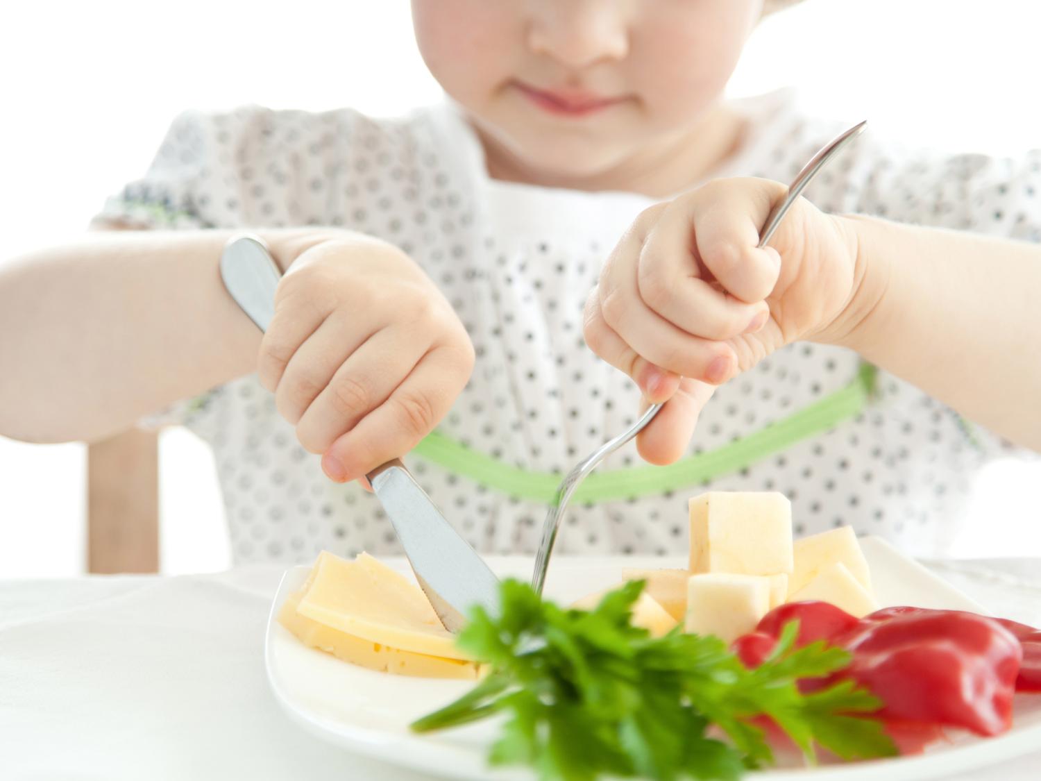 A young child using a knife and fork to eat a plate of vegetables