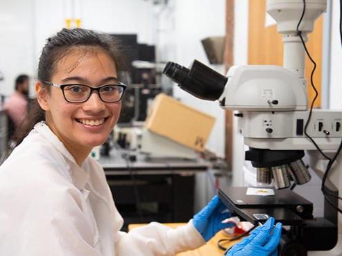 A researcher smiles at the camera while sitting in front of a microscope
