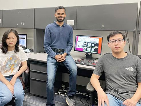 Three people smile at the camera in front of a computer monitor displaying graphs 