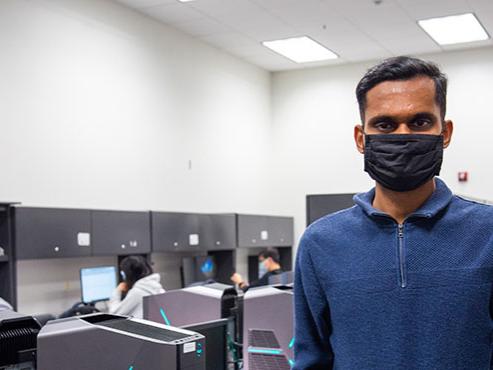 A man in a blue long-sleeve shirt and a black face mask faces the camera in front of a computer lab.