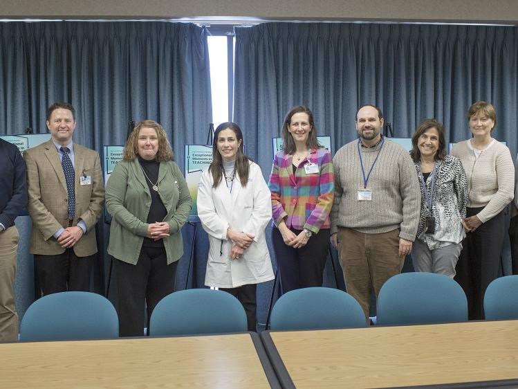Nine women and men stand in a row front of easels and drawn curtains, behind chairs positioned at a table.