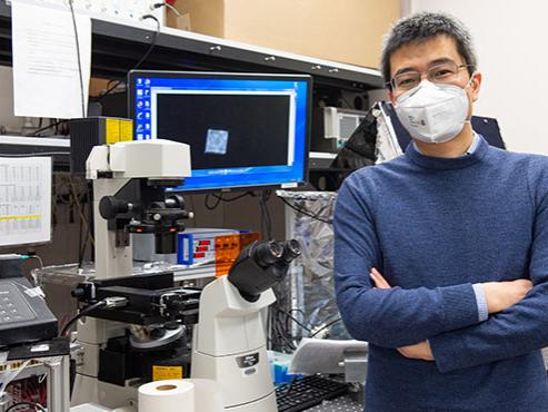A man wearing glasses, a mask and a blue sweater poses with his arms crossed in front of a computer screen and microscope. 