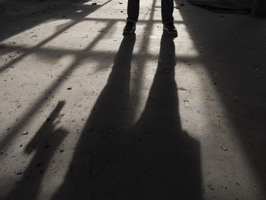 A person casts a shadow on the pavement. The photo focuses on the person’s shoes, and their shadow reveals that they are holding a gun. 