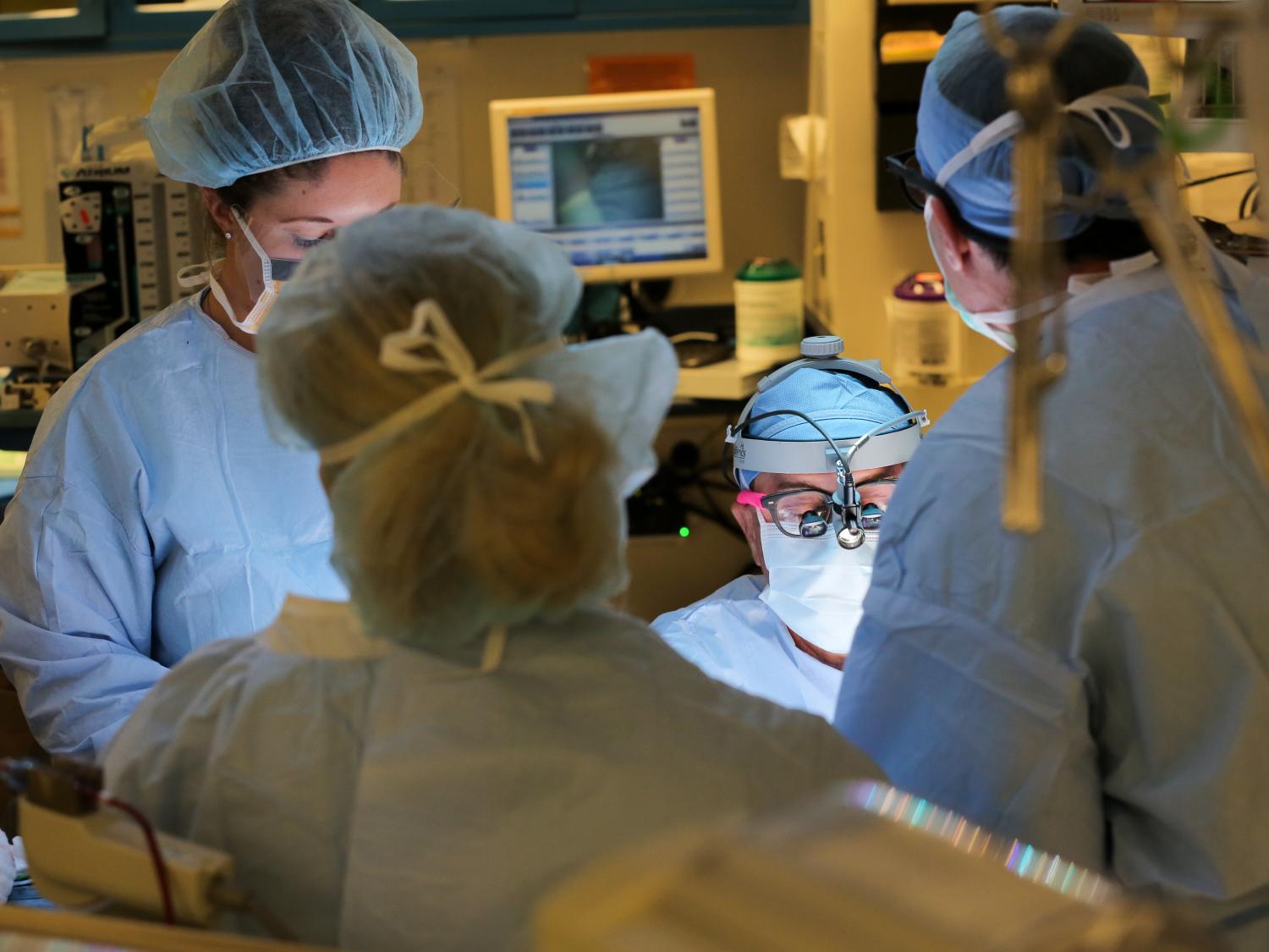 Four people in surgical scrubs and gowns are gathered around a patient in the operating room. A computer monitor and some equipment are visible in the background.