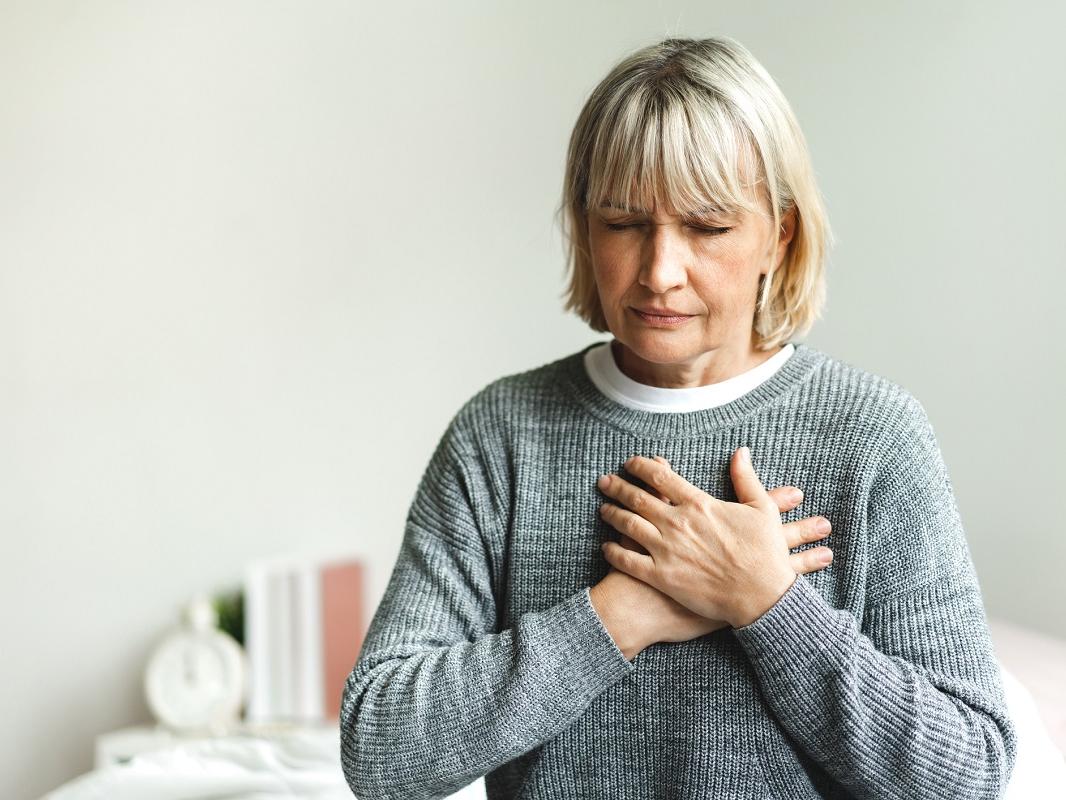 A woman sits on the edge of a bed with chest pain, her hands placed over her chest.