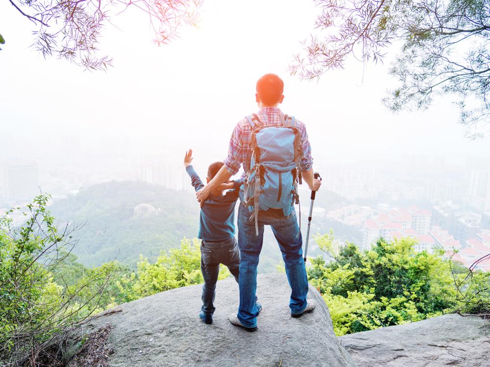 The view from behind as a man and child, dressed in hiking gear, stand on a rock looking off the edge of a mountain. Trees are in the foreground. Buildings and more trees and hills are in the background.