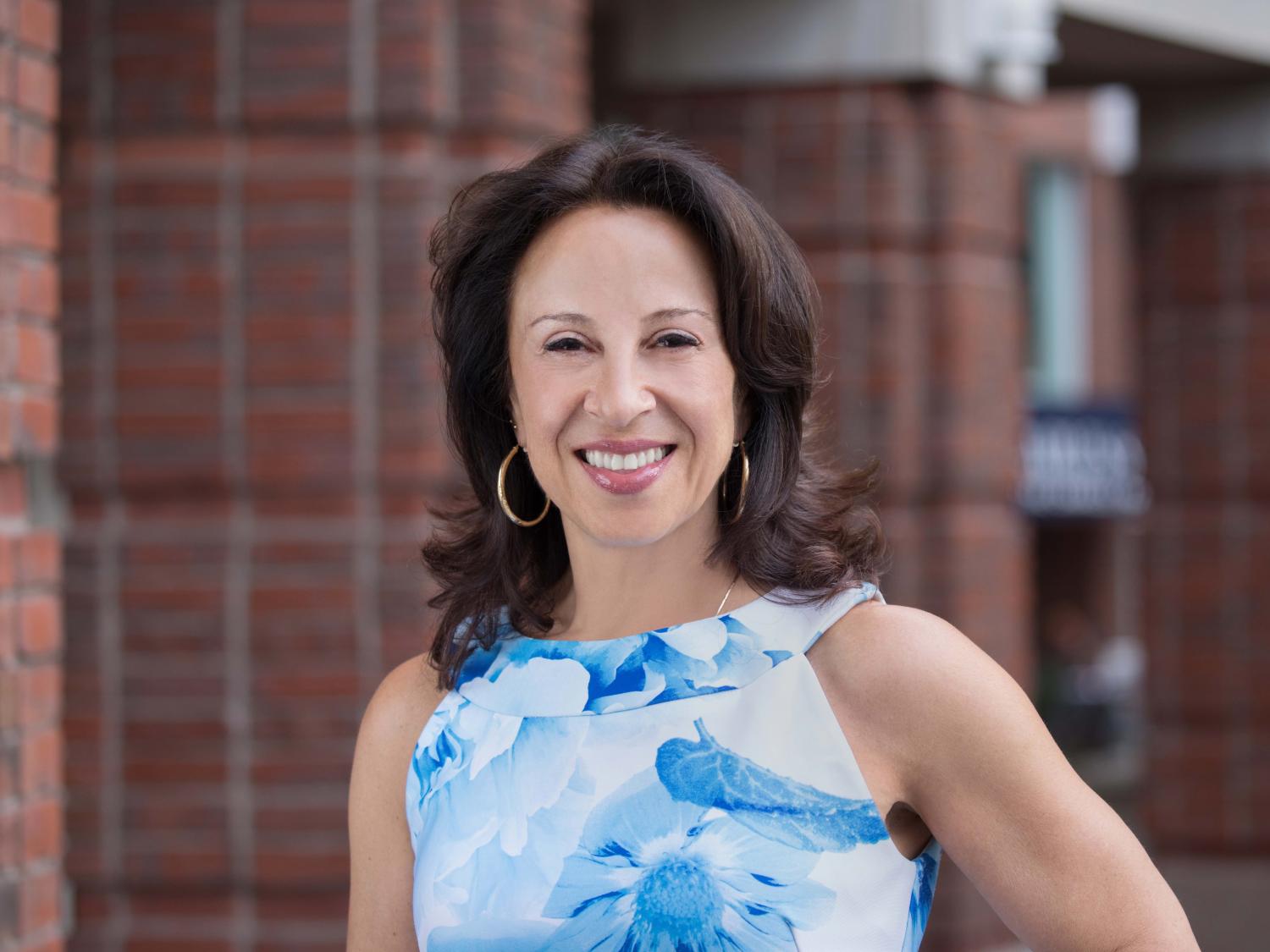 Maria Hinojosa wears a blue and white top and smiles at the camera in front of a brick structure
