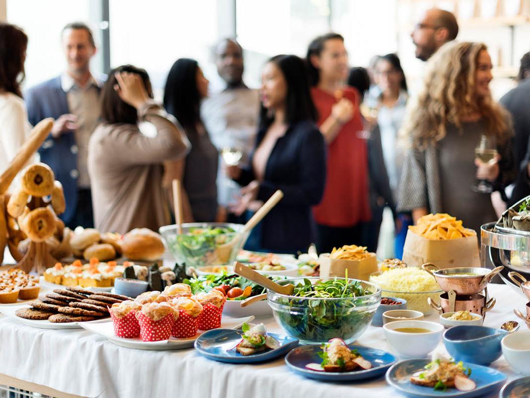 A table with plates and bowls of food is in the foreground. It includes a salad, cookies, muffins, bread and various hors d’oeuvres. In the background, several people are standing and talking, some of them holding drinks.
