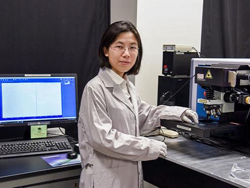 A woman in a lab coat and gloves holds a sample under a microscope in a lab while looking at the camera.