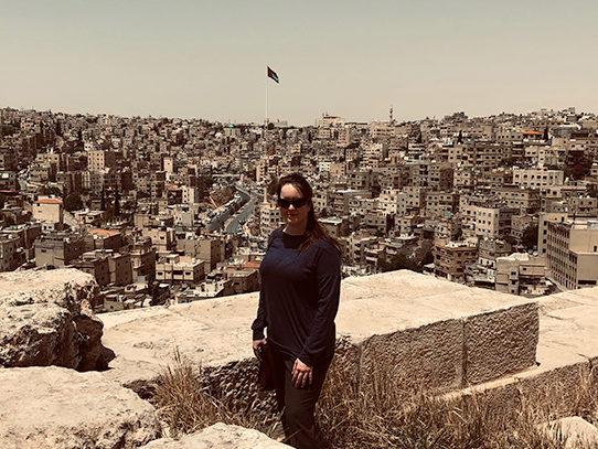 A woman wearing dark clothing stands at an overlook of a city with hundreds of buildings behind her.