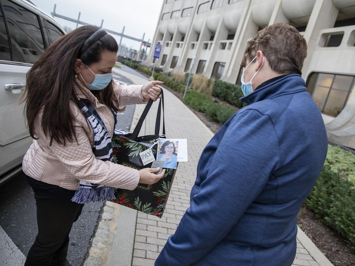 A woman in a mask stands next to a sports utility vehicle and shows someone else in a mask the contents of a bag, which includes the photo of a woman.