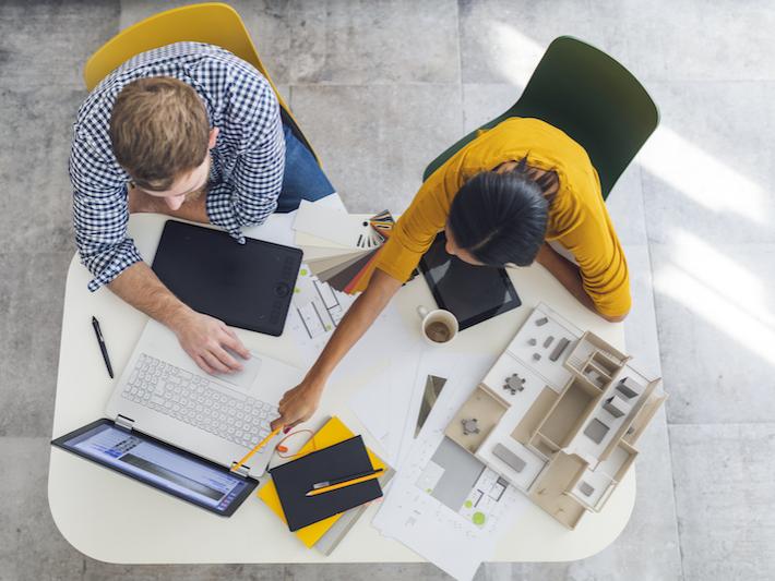 Overhead photo of two people sitting at a desk with a laptop, papers and a model of a house. One person is pointing at the laptop